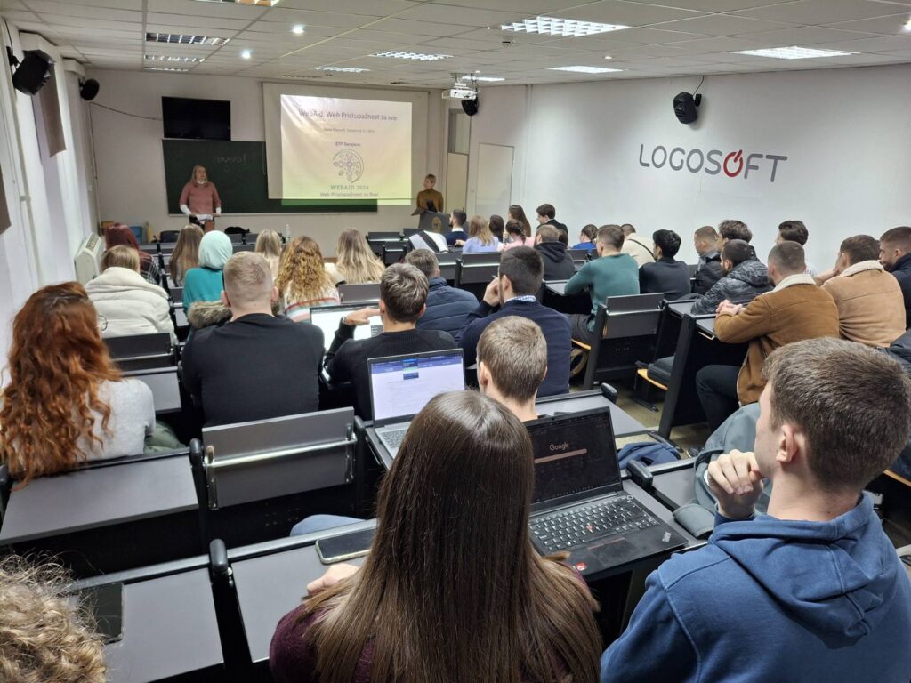 Studenti u prilično punoj predavaonici slušaju, predavačica predaje, projekcija na platnu