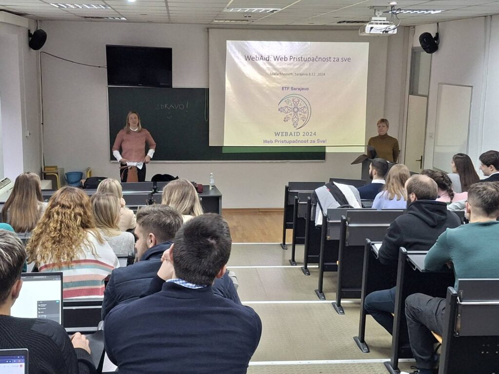 Studenti u prilično punoj predavaonici slušaju, predavačica predaje, projekcija na platnu